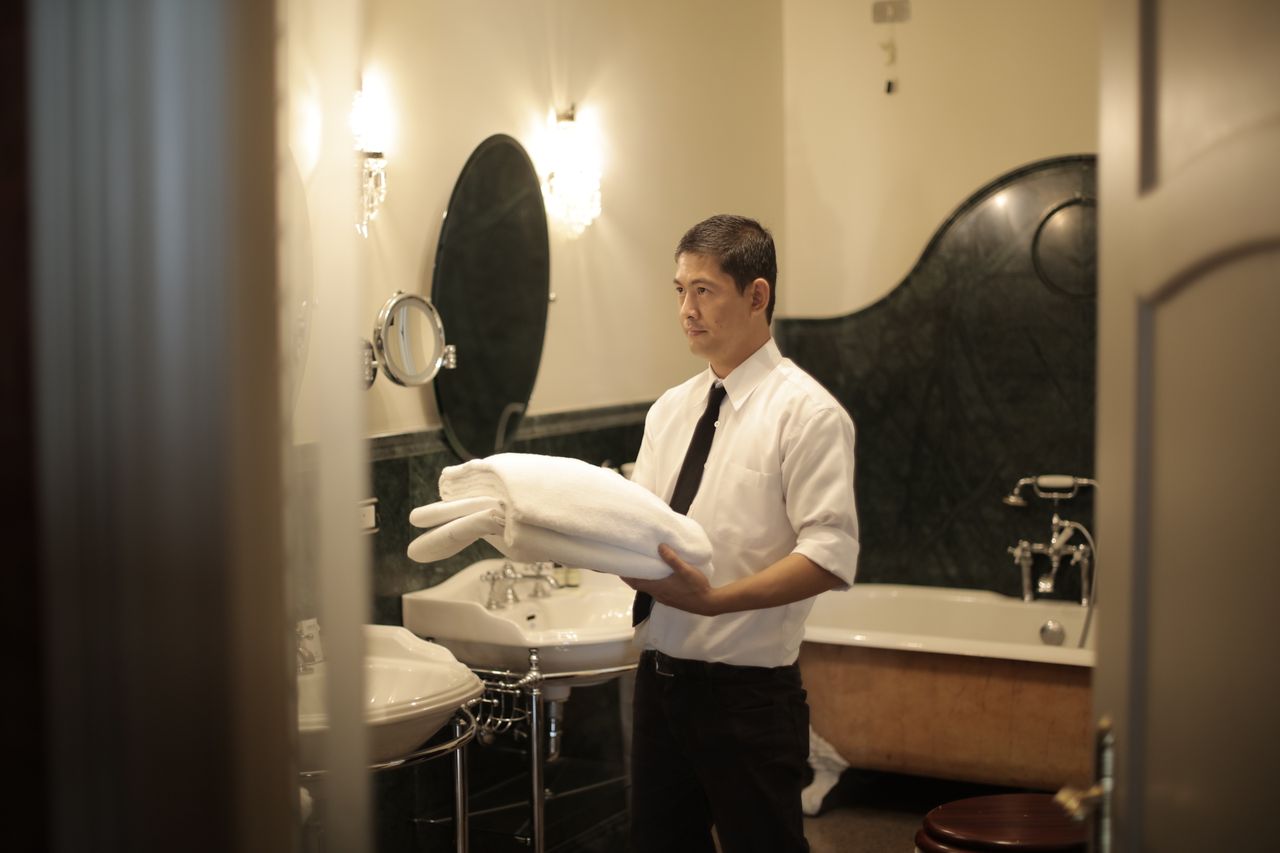 about-img-01 Side view of young male employee holding stack of white towels while standing in bathroom of luxury hotel